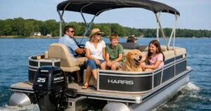 A family of four sits together on a pontoon boat cruising across a calm lake on a sunny day. An adult steers the boat while the others relax on cushioned seats, accompanied by a golden retriever sitting happily at the front. Trees and lakeside homes are visible in the background, creating a peaceful recreational setting.